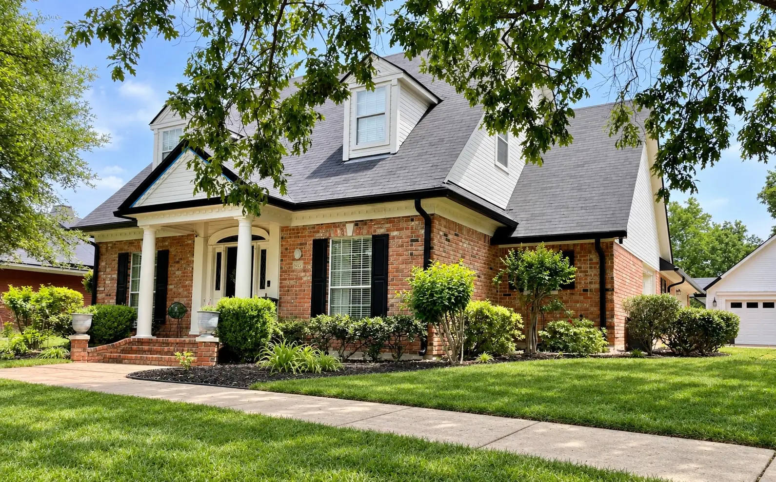 Brick home with dormers, dark gutters, white porch columns and an arched entry overlooking a tidy front garden
