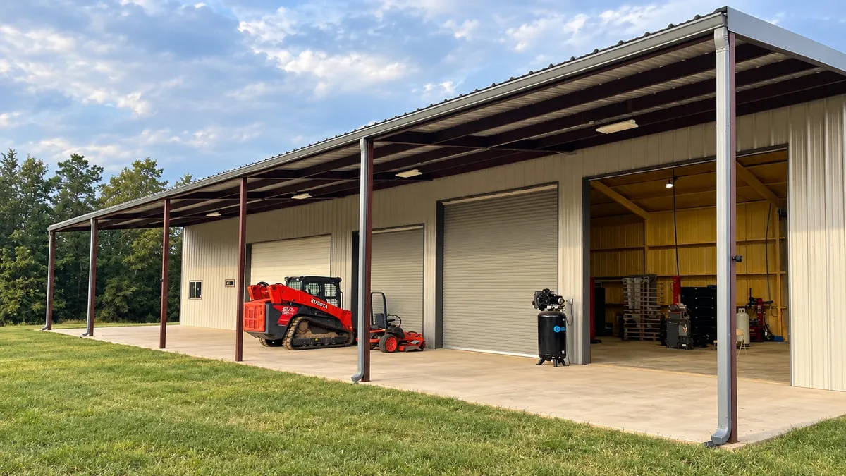 Commercial metal building with gutters and downspouts along a long covered bay, red work equipment, and a tidy concrete pad bordered by grass