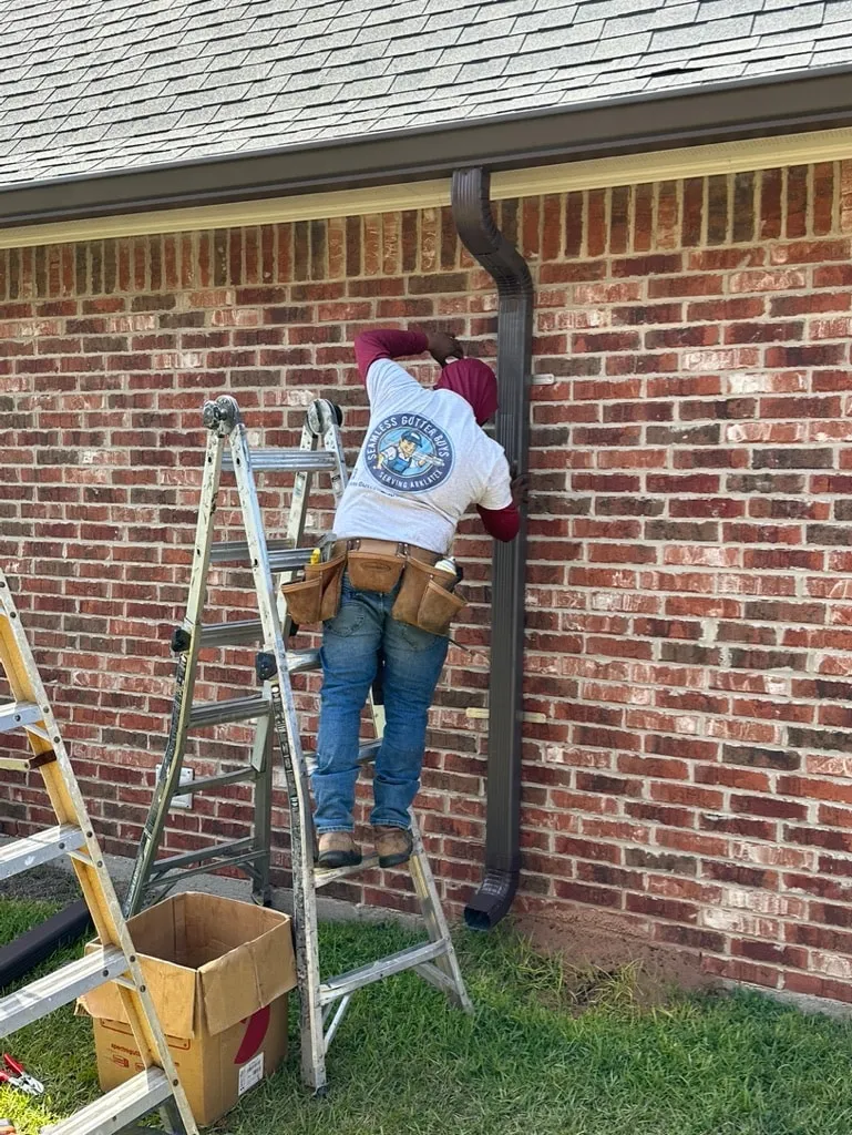 Seamless Gutter Guys crew member on a ladder working on a dark downspout against a red brick wall