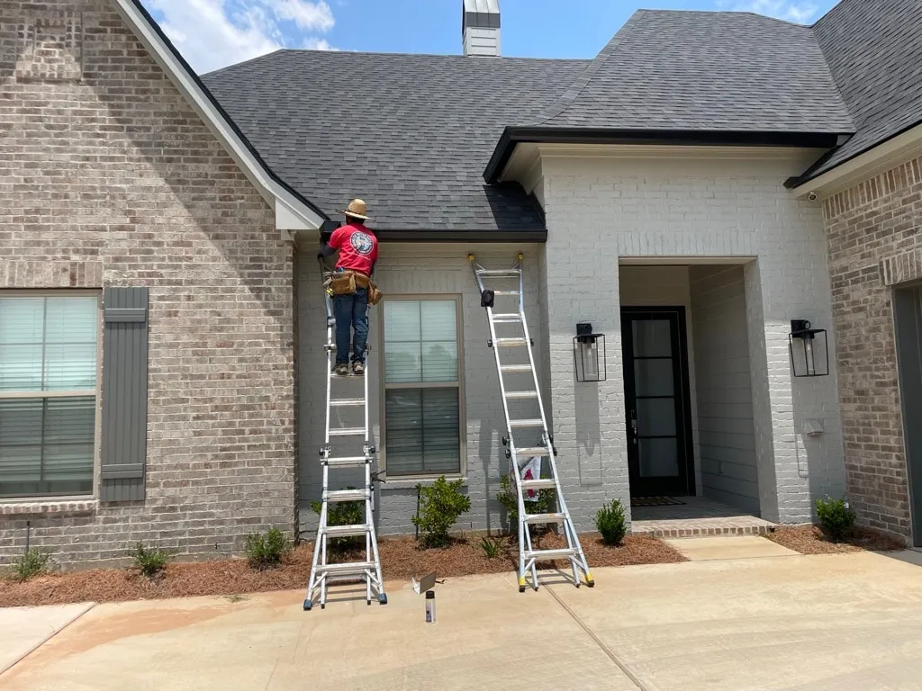 Seamless Gutter Guys installer in a red company shirt working on a dark gutter above the entry courtyard of a brick home