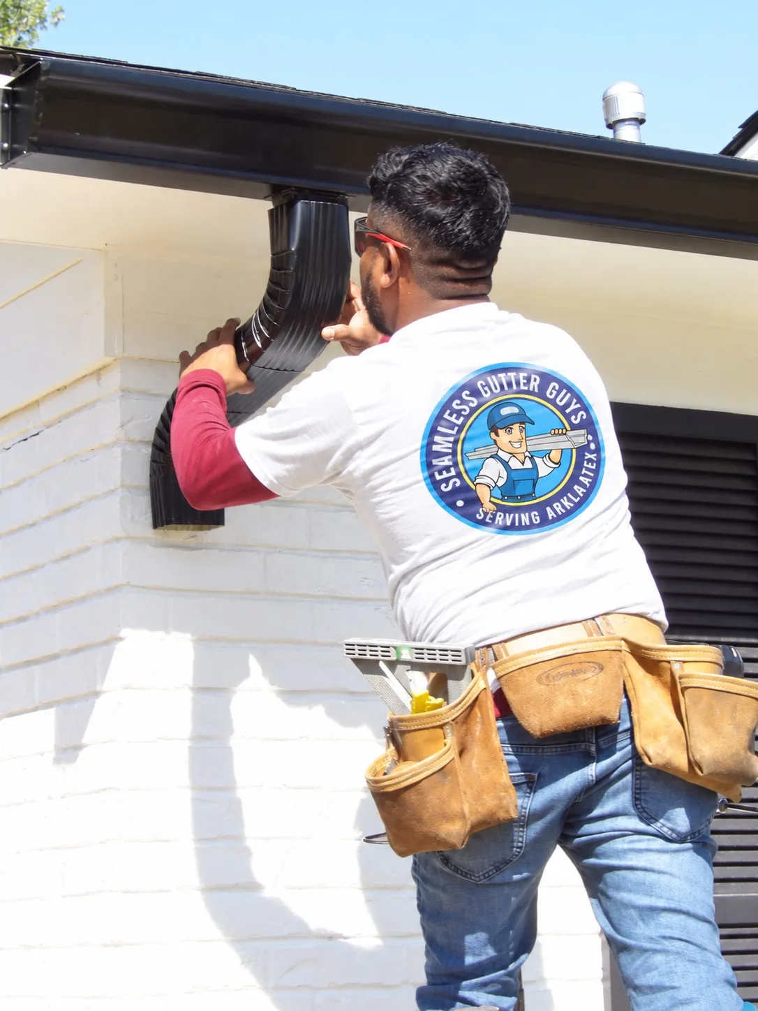 Seamless Gutter Guys technician in a white company shirt working on a dark downspout elbow at a white brick home