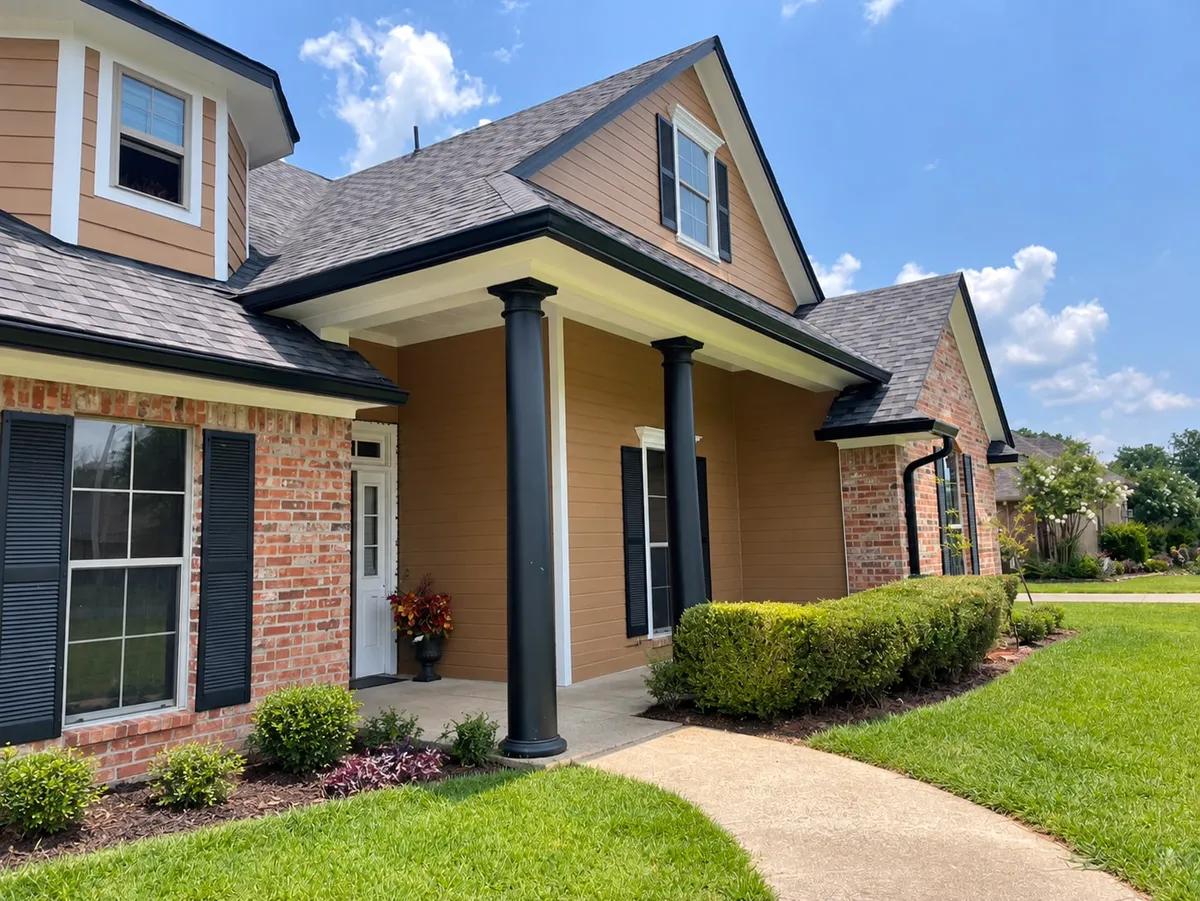 Tan-sided and brick home with black gutters and a downspout, dark porch columns, trimmed hedges and a clear front walkway