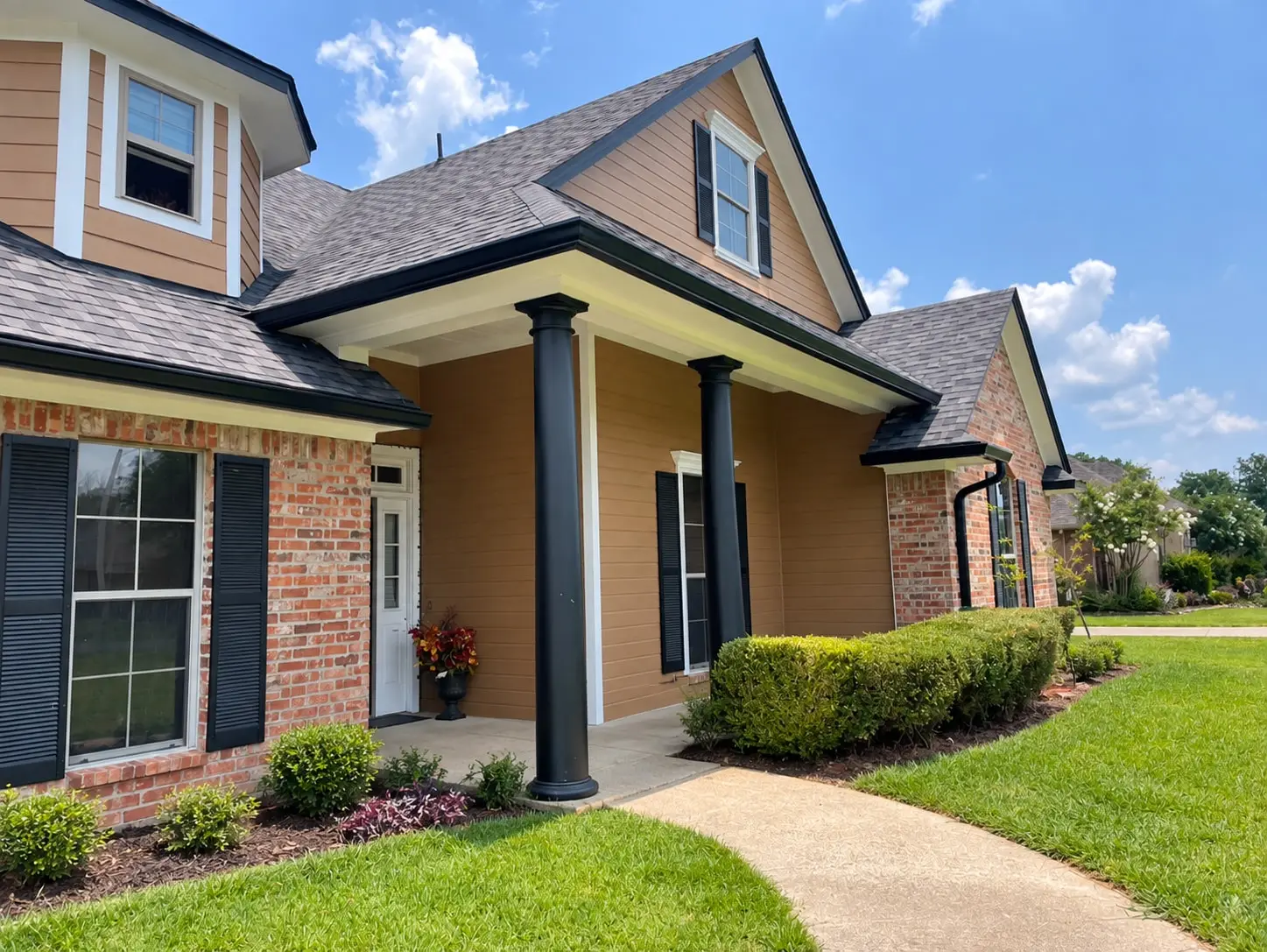 Tan-sided and brick home with black gutters and a downspout, dark porch columns, trimmed hedges and a clear front walkway