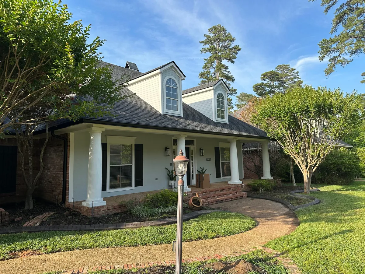 White cottage with dormer windows, porch columns and dark gutters, behind a curved brick walkway